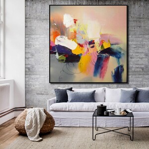 May include: Abstract painting with bold strokes of yellow, pink, blue, and green on a light gray background. The painting is framed in black and hanging on a concrete wall above a white sofa.
