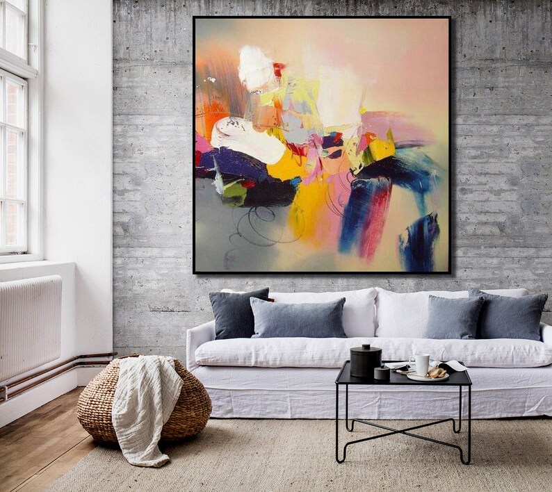 May include: Abstract painting with bold strokes of yellow, pink, blue, and green on a light gray background. The painting is framed in black and hanging on a concrete wall above a white sofa.