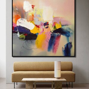 May include: Abstract painting with bold strokes of yellow, pink, blue, and white paint on a canvas. The painting is framed in black and hangs on a white wall above a brown sofa and a coffee table.
