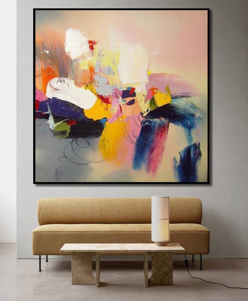 May include: Abstract painting with bold strokes of yellow, pink, blue, and white paint on a canvas. The painting is framed in black and hangs on a white wall above a brown sofa and a coffee table.