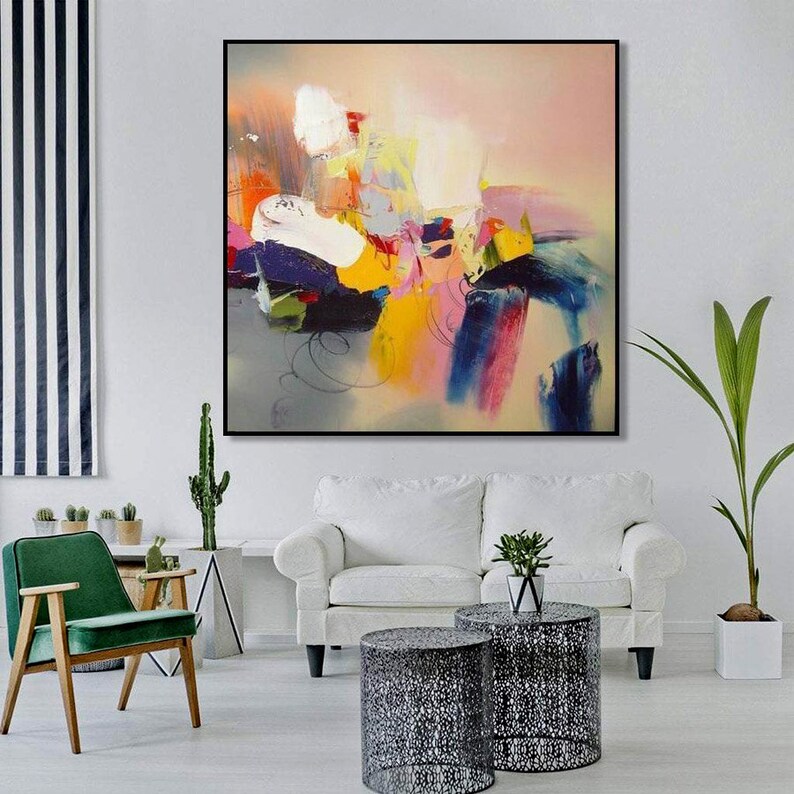 May include: Abstract painting with bold strokes of pink, yellow, blue, and green on a light gray background. The painting is framed in black and hanging on a white wall.