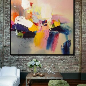 May include: Abstract painting with bold strokes of yellow, blue, pink, and white on a canvas. The painting is framed and hanging on a brick wall.