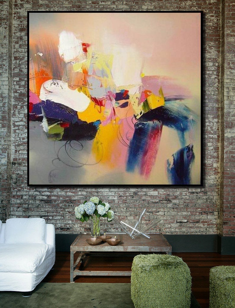 May include: Abstract painting with bold strokes of yellow, blue, pink, and white on a canvas. The painting is framed and hanging on a brick wall.