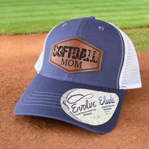 Peut inclure: Une casquette de baseball bleue et blanche avec un patch en cuir marron qui dit "Softball Mom". La casquette a un dos en mesh et une visière blanche.