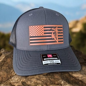 May include: A gray and black trucker hat with an American flag and a silhouette of a lineman climbing a utility pole. The hat has a brown leather patch with the text "EST. 1970 RICHARDSON. ORIGINAL 112 TRUCKER ADJUSTABLE".