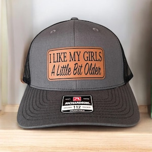 Puede incluir: Una gorra de camionero gris y negra con un parche de cuero marrón que dice "ME GUSTAN MIS CHICAS UN POCO MÁS MAYORES". La gorra es ajustable y tiene el logotipo de Richardson 112.
