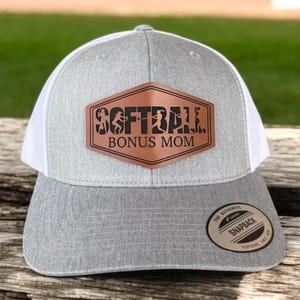 Op de afbeelding: Grijze en witte truckercap met een bruin leren patch waarop "Softball Bonus Mom" staat. De pet heeft een wit mesh achterpaneel en een bruin "The Authentic Snapback" label.