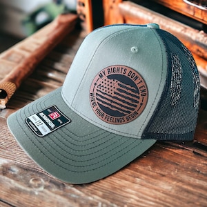 May include: A gray and navy blue trucker hat with a circular patch featuring an American flag design and the text "MY RIGHTS DON'T END WHERE YOUR FEELINGS BEGIN." The hat has a curved brim and a mesh back.
