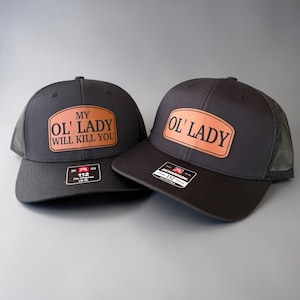 May include: Two black trucker hats with a brown leather patch. One hat reads "MY OL' LADY WILL KILL YOU" and the other reads "OL' LADY". Both hats have a mesh back and a curved brim.