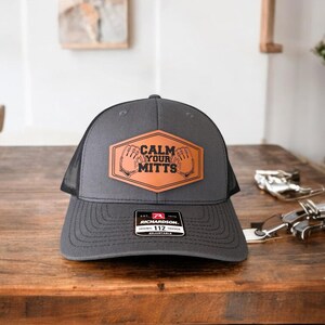 Könnte beinhalten: Eine schwarze und graue Trucker-Kappe mit einem braunen Leder-Patch, auf dem "Calm Your Mitts" steht. Die Kappe hat ein schwarzes Netzrücken und einen verstellbaren Richardson 112-Riemen.