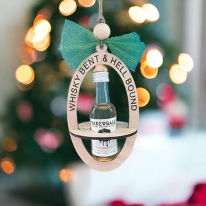 May include: A wooden Christmas ornament shaped like an oval with the text "Whiskey Bent & Hell Bound" around the edge. A miniature bottle of "Skrewball Peanut Butter Whiskey" is inside the ornament.
