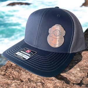 May include: A navy blue and gray trucker hat with a silver metal diving helmet patch. The hat has a black adjustable strap with the text "Richardson 112 Original Trucker Adjustable" on it.