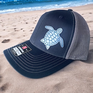 Pode incluir: Um boné de caminhoneiro azul marinho e cinza com um gráfico de tartaruga marinha azul claro. O boné tem uma parte de trás de malha e uma aba curva. O boné está em uma praia de areia.