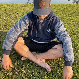 May include: A person wearing a dark blue hoodie with camouflage sleeves and a dark gray baseball cap with a brown leather patch that says "Support Your Local Pole Dawg". The person is sitting cross-legged on a grassy surface.
