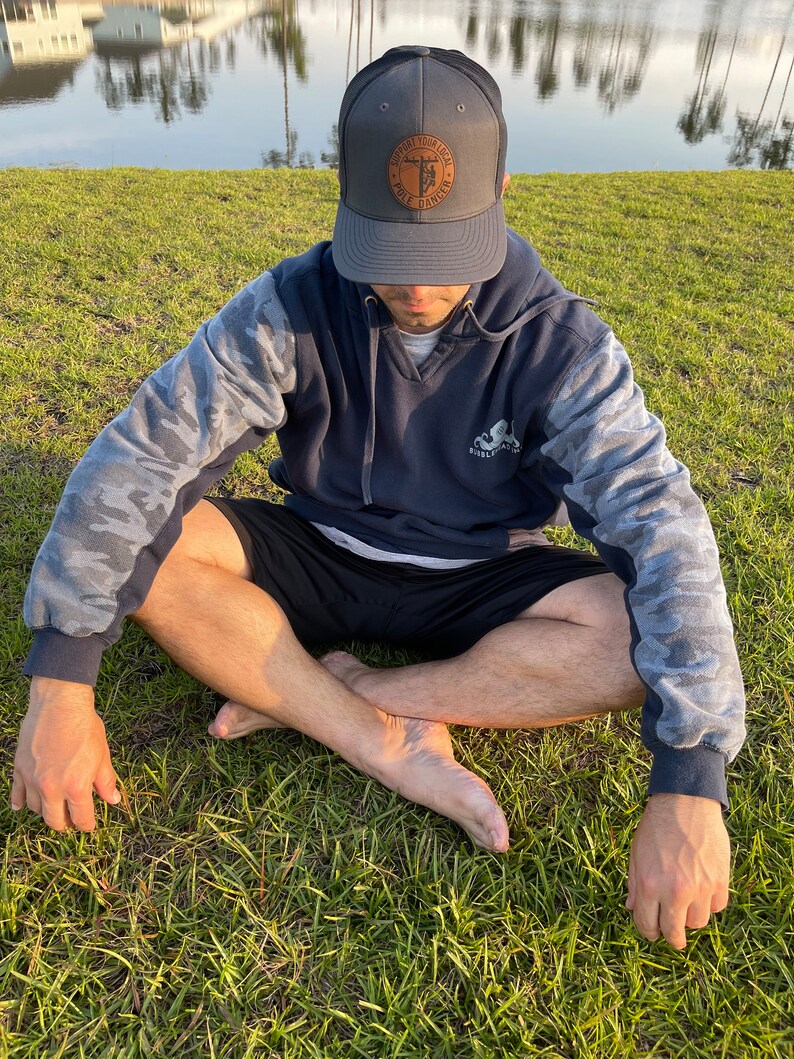 May include: A person wearing a dark blue hoodie with camouflage sleeves and a dark gray baseball cap with a brown leather patch that says "Support Your Local Pole Dawg". The person is sitting cross-legged on a grassy surface.