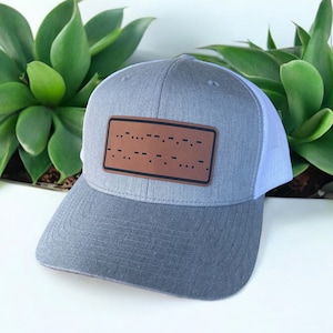 Könnte beinhalten: Eine graue und weiße Trucker-Kappe mit einem braunen Leder-Patch mit Braille-Punkten. Der Patch lautet "..."