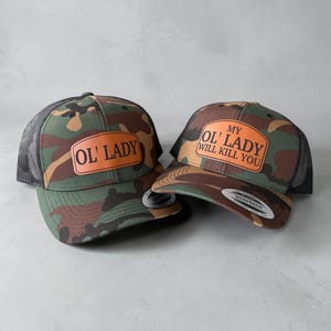 Puede incluir: Dos gorras de camionero de camuflaje con la parte trasera de malla negra. Una gorra tiene el texto "OL' LADY" en un parche naranja. La otra gorra tiene el texto "MY OL' LADY WILL KILL YOU" en un parche naranja. Ambas gorras tienen una visera curva.