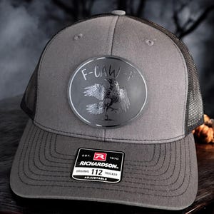 May include: Gray trucker hat with a black mesh back and a curved brim. The hat features a circular patch with a crow illustration and the text "F-CAW-F". A black and white Richardson patch is on the front.