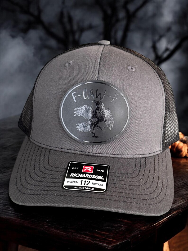 May include: Gray trucker hat with a black mesh back and a curved brim. The hat features a circular patch with a crow illustration and the text "F-CAW-F". A black and white Richardson patch is on the front.