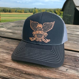 Può includere: Un cappello da camionista nero e grigio con una toppa in pelle marrone che presenta un'aquila con il testo "We the People Shall Not Be Infringed".