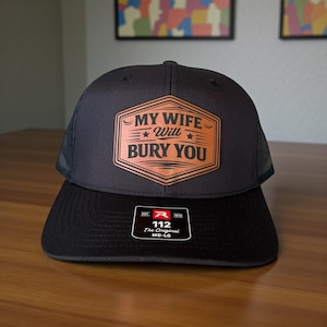 May include: Black and gray trucker hat with a brown patch that reads "MY WIFE Will BURY YOU". The hat has a mesh back and a curved brim. The hat has a tag that reads "112 The Original MD-LG".