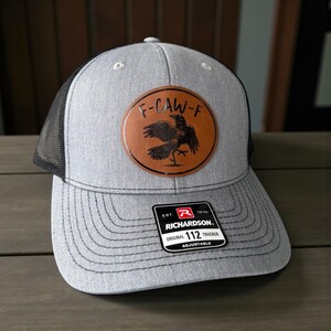 May include: Gray trucker hat with a black mesh back and a brown leather patch. The patch features a crow illustration and the text "F-CAW-F". The hat has a curved brim and an adjustable strap. The tag reads "RICHARDSON 112 TRUCKER".
