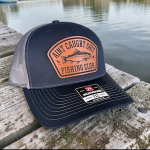 Peut inclure: Casquette de camionneur bleu marine et grise avec un écusson en cuir portant l'inscription "Aint Caught Shit Fishing Club" et un dessin de poisson. La casquette est réglable et porte une étiquette Richardson 112.