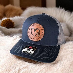 May include: A navy blue and gray trucker hat with a brown leather patch that says "Motivated by Caffeine & Canines" with a paw print and heart design. The hat is made by Richardson 112.