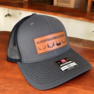 Pu&ograve; includere: Un cappello da camionista grigio e nero con una toppa in pelle marrone che dice "#ILIKEMYRACKSBIGANDFIRM" con un grafico di corna di cervo e reggiseno. Il cappello &egrave; regolabile e ha un'etichetta Richardson 112.
