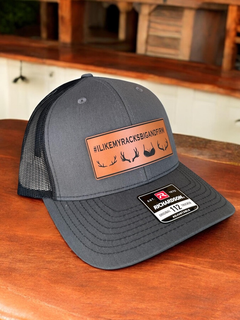 Pu&ograve; includere: Un cappello da camionista grigio e nero con una toppa in pelle marrone che dice "#ILIKEMYRACKSBIGANDFIRM" con un grafico di corna di cervo e reggiseno. Il cappello &egrave; regolabile e ha un'etichetta Richardson 112.