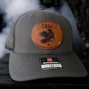 May include: Gray trucker hat with a mesh back and a curved brim. The hat features a brown leather patch with a raven design and the text "F-CAW-F". The Richardson 112 Trucker hat is adjustable.