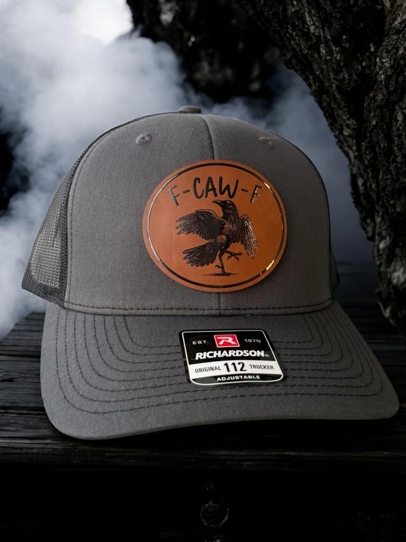 May include: Gray trucker hat with a mesh back and a curved brim. The hat features a brown leather patch with a raven design and the text "F-CAW-F". The Richardson 112 Trucker hat is adjustable.