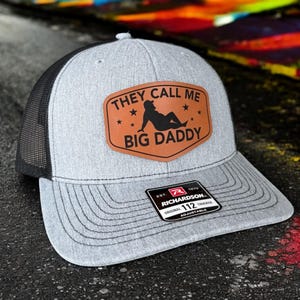 Op de afbeelding: Grijze truckerpet met een zwarte mesh achterkant en een gebogen klep. De pet heeft een bruin leren patch met de tekst "THEY CALL ME BIG DADDY" en een silhouet. De pet is verstelbaar en heeft een Richardson-label.