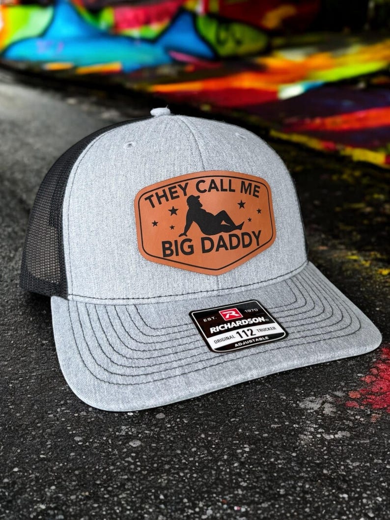 Op de afbeelding: Grijze truckerpet met een zwarte mesh achterkant en een gebogen klep. De pet heeft een bruin leren patch met de tekst "THEY CALL ME BIG DADDY" en een silhouet. De pet is verstelbaar en heeft een Richardson-label.