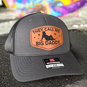 Op de afbeelding: Grijze truckerpet met een zwarte mesh achterkant en een gebogen klep. De pet heeft een bruine patch met de tekst "THEY CALL ME BIG DADDY" en een silhouet-afbeelding. De pet is verstelbaar.