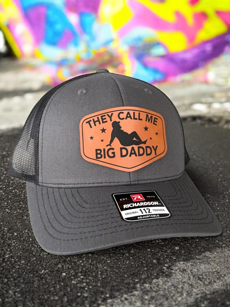 Op de afbeelding: Grijze truckerpet met een zwarte mesh achterkant en een gebogen klep. De pet heeft een bruine patch met de tekst "THEY CALL ME BIG DADDY" en een silhouet-afbeelding. De pet is verstelbaar.