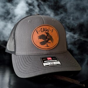 May include: Gray trucker hat with a black mesh back and a leather patch. The patch features a raven design with the text "F-CAW-F". The hat has a Richardson 112 tag.