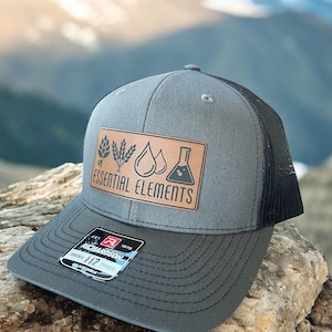 May include: A grey trucker hat with a black mesh back. The hat features a brown leather patch with the words "Essential Elements" and graphic icons. The hat has a curved brim and a Richardson 112 sticker.