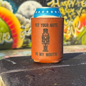Puede incluir: Un enfriador de latas de cuero sintético marrón con el texto "PUT YOUR NUTS IN MY MOUTH" y un gráfico de cascanueces. Una lata azul y blanca está dentro del enfriador. El fondo es una colorida pared de graffiti.