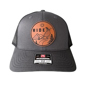 Peut inclure: Casquette de camionneur grise avec un dos en filet noir et une visière incurvée. Le chapeau présente un écusson circulaire avec le mot "RIDE" et une scène de VTT. L'écusson est marron avec un contour noir. Le chapeau a un écusson Richardson.