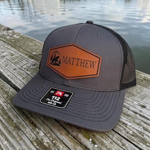 May include: Grey trucker cap with a black mesh back and a brown leather patch. The patch features a bear and mountain graphic with the name "MATTHEW". The cap has a curved peak and a tag that reads "112 The Original MD-LG".