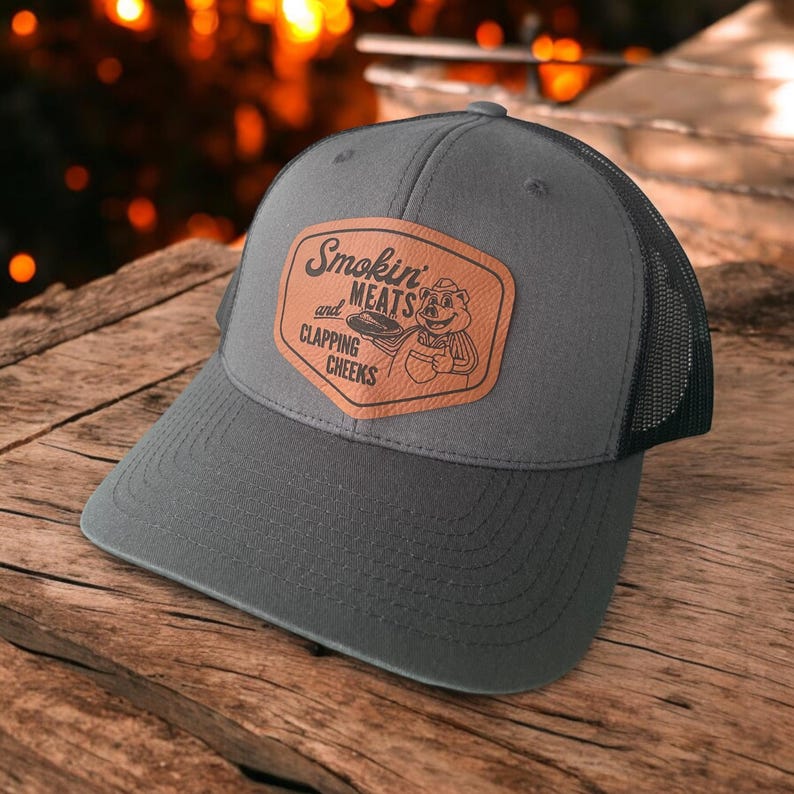 May include: A gray trucker hat with a mesh back and a curved brim. The hat features a brown leather patch with the words "Smokin' Meats and Clapping Cheeks" and a cartoon pig graphic. The hat is on a wooden surface.