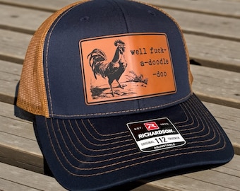 May include: A navy blue trucker hat with a brown mesh back and a brown leather patch on the front. The patch features a black illustration of a rooster and the text well fuck-a-doodle-doo. A black Richardson 112 adjustable sticker is on the brim.