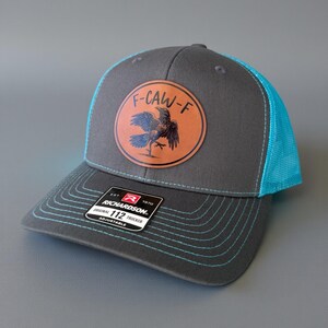 May include: A gray trucker hat with a teal mesh back and brim stitching. The hat features a circular patch with a raven illustration and the text "F-CAW-F". The Richardson 112 Trucker hat is adjustable.