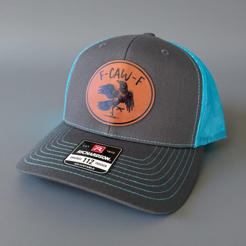 May include: A gray trucker hat with a teal mesh back and brim stitching. The hat features a circular patch with a raven illustration and the text "F-CAW-F". The Richardson 112 Trucker hat is adjustable.