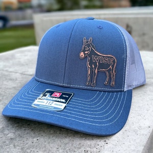 Könnte beinhalten: Eine blaue und graue Trucker-Kappe mit einem rosafarbenen Esel-Motiv und dem Wort "WISE" auf der Vorderseite. Die Kappe ist verstellbar und hat ein Richardson 112-Etikett.