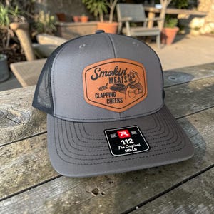 May include: A gray trucker hat with a black mesh back and a curved brim. The front features a brown leather patch with the words "Smokin' Meats and Clapping Cheeks" and a cartoon pig. A tag on the brim reads "112 The Original MD-LG".