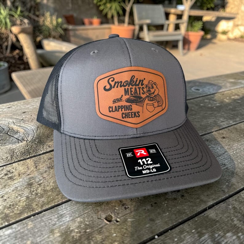 May include: A gray trucker hat with a black mesh back and a curved brim. The front features a brown leather patch with the words "Smokin' Meats and Clapping Cheeks" and a cartoon pig. A tag on the brim reads "112 The Original MD-LG".