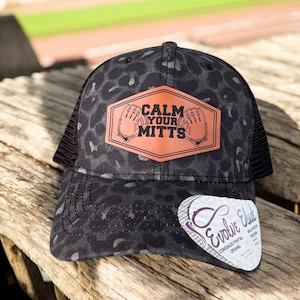 May include: Black and white leopard print trucker hat with a brown leather patch that says "Calm Your Mitts".