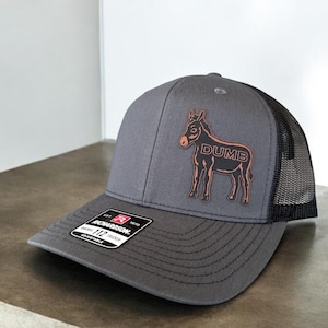 May include: A gray trucker hat with a black mesh back. The hat features a brown donkey graphic with the word "DUMB" in brown lettering. The hat has a curved brim and an adjustable snap closure.